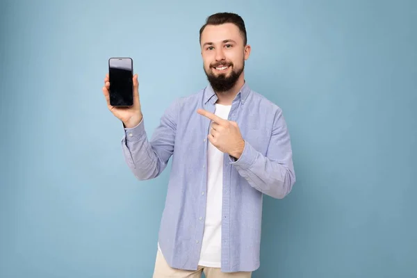 Foto de bonito sorridente adulto masculino pessoa bonita vestindo roupas casuais em pé isolado no fundo com espaço de cópia segurando smartphone mostrando telefone na mão com tela vazia para — Fotografia de Stock