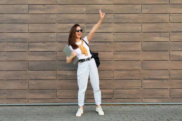 Full length photo shot of young charming beautifyl brunet woman going in the street near brown wall looking to side and showing peace gesture in white t-shirt and light blue jeans white sneakers with — Stock Photo, Image