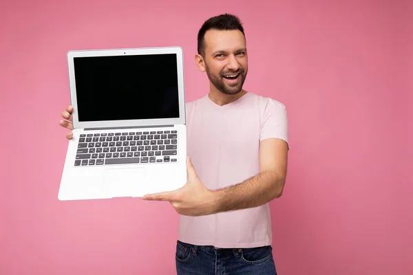 Guapo sonriente morena hombre sosteniendo ordenador portátil mirando a la cámara en camiseta sobre fondo rosa aislado —  Fotos de Stock