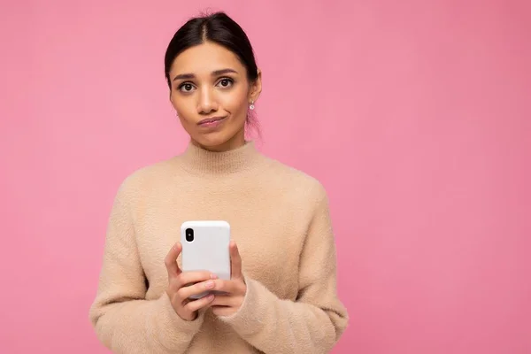 Wanita muda berambut coklat yang lucu dan menarik, mengenakan sweater hangat berwarna krem, berdiri terisolasi di atas latar belakang merah muda berselancar di internet melalui telepon sambil melihat kamera. — Stok Foto