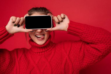Kırmızı arka plan duvarında izole edilmiş sıcak kırmızı süveter giyen güzel, tatlı, gülümseyen genç bir kadının fotoğrafı. Elinde akıllı telefon var ve telefonu boş bir şekilde gösteriyor.