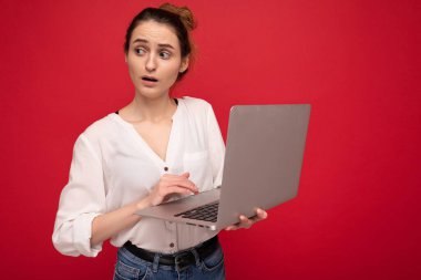 Netbook bilgisayarını elinde tutan, ağzı açık beyaz gömlekli, kırmızı arka planda izole edilmiş beyaz gömlekli güzel esmer kadının yan profili.