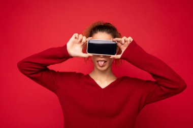 Kırmızı arka plan üzerinde koyu kırmızı süveter giyen mutlu genç bir kadının fotoğrafı. Akıllı telefonu tutuyor ve cep telefonu ekranını boş bir şekilde gösteriyor.