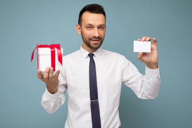 Mavi arka plan duvarında sakallı, sakallı, yakışıklı, mutlu, esmer bir adamın beyaz gömlek ve kravat takıp kırmızı kurdeleli beyaz bir hediye kutusu tutarken çekilmiş fotoğrafı.