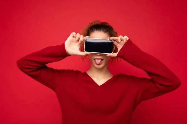 Foto retrato de feliz engraçado jovem mulher vestindo suéter vermelho escuro isolado sobre fundo vermelho segurando smartphone e mostrando tela do telefone móvel com espaço vazio para mockup se divertindo — Fotografia de Stock