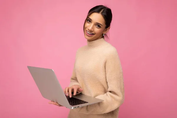 Fotografie de frumos zâmbitor fericit tânără brunetă femeie purtând pulover bej care deține netbook calculator tastarea text pe tastatură uitându-se la camera izolată peste fundal perete roz — Fotografie, imagine de stoc
