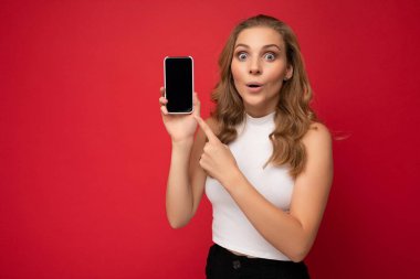 Şaşırdım. Beyaz tişört giyen güzel sarışın kadın kırmızı arka planda izole edilmiş. Fotokopi alanı elinde akıllı telefonuyla elinde boş bir ekranla kameraya bakıyor.