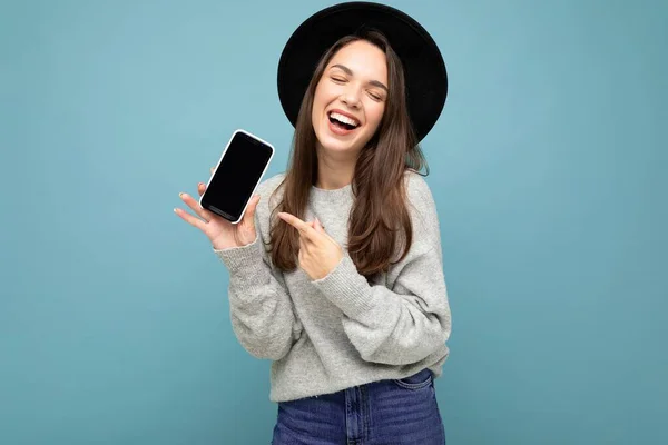 검은 모자와 회색 스웨터를 입고 배경 화면에 분리 된 전화기를 가리키는 손가락을 들고 있는 매력적 인 젊고 행복 한 여성의 모습을 클로즈업 — 스톡 사진