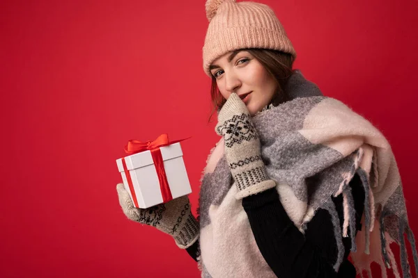 Shot of charming sexy positive young brunette woman isolated over red background wall wearing winter scarf hat and mittens holding white gift box with red ribbon and looking at camera and keeping