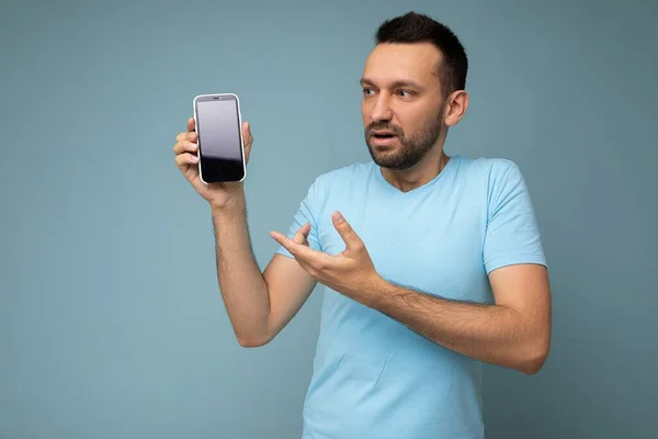 Sorprendido joven guapo sin afeitar morena hombre usando todos los días camiseta azul aislado sobre fondo azul celebración y mostrando el teléfono móvil con pantalla vacía para recortar mirando el teléfono inteligente —  Fotos de Stock