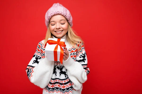 Foto av vacker glad leende ung blond kvinna isolerad över röd bakgrund vägg bär vinter tröja och rosa hatt innehav vit presentförpackning med rött band och tittar på nuläget — Stockfoto