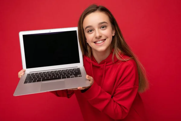 Foto de retrato de cerca de la hermosa chica sonriente feliz con el pelo largo con capucha roja que sostiene el ordenador portátil mirando a la cámara aislada sobre el fondo de la pared roja —  Fotos de Stock