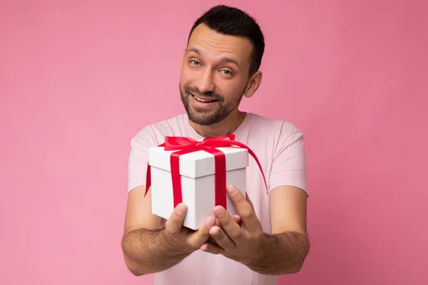 Foto de guapo morena positiva sin afeitar joven aislado sobre la pared de fondo rosa con camiseta rosa sosteniendo caja de regalo blanca con cinta roja y mirando a la cámara —  Fotos de Stock