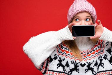 Çekici, sarışın, kalın örgü şapkalı ve kalın kazaklı genç bir kadının fotoğrafı. Kırmızı arka planda izole bir şekilde duruyor. Akıllı telefonlu, boş ekranlı kameraya bakıyor.