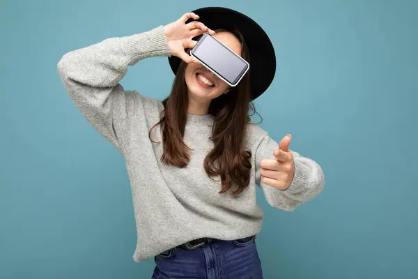 Fechar-se de atraente jovem feliz mulher vestindo chapéu preto e camisola cinza segurando dedo apontando telefone para a câmera isolada no fundo — Fotografia de Stock