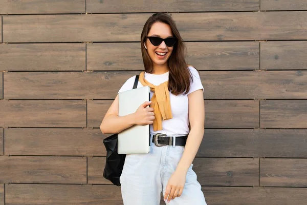 Beautiful smiling happy charming young brunet woman looking at camera holding computer laptop and black sunglasses in white t-shirt and light blue jeans in the street near brown wall — Stock Photo, Image