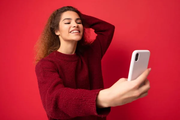 Attractive charming young smiling happy woman holding and using mobile phone taking selfie wearing stylish clothes isolated over wall background — Stock Photo, Image