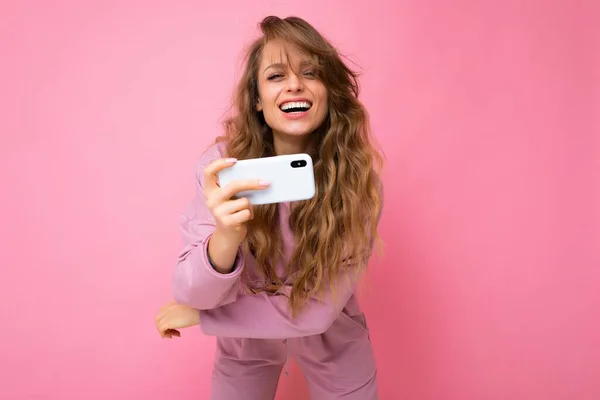 Tiros de alegria sorrindo feliz Linda jovem loira vestindo capuz rosa isolado sobre fundo rosa com espaço de cópia segurando e usando telefone celular olhando para a câmera — Fotografia de Stock