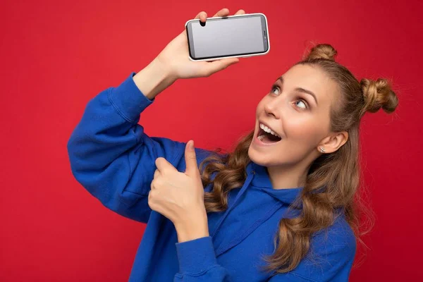 Atraente chocado sorrindo jovem loira vestindo elegante capuz azul isolado no fundo vermelho com espaço de cópia segurando smartphone mostrando telefone na mão com tela vazia apontando para — Fotografia de Stock