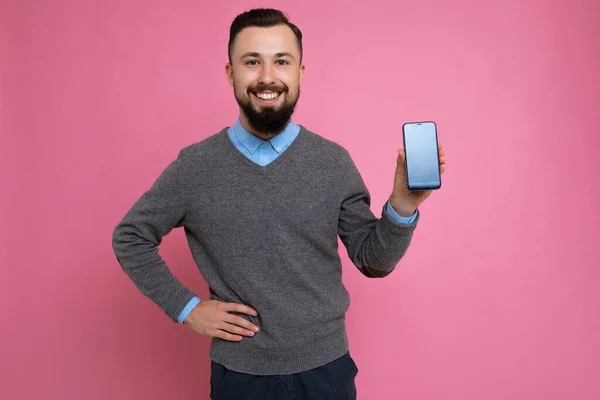 Positive handsome happy cool young brunette unshaven man with beardwearing stylish grey sweater and blue shirt standing isolated over pink background wall holding smartphone and showing phone with — ストック写真