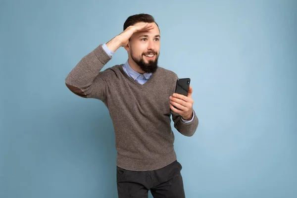 Closeup portrait of Handsome smiling brunette bearded man wearing grey sweater and blue shirt isolated on background wall holding smartphone looking to the side — Stock Photo, Image