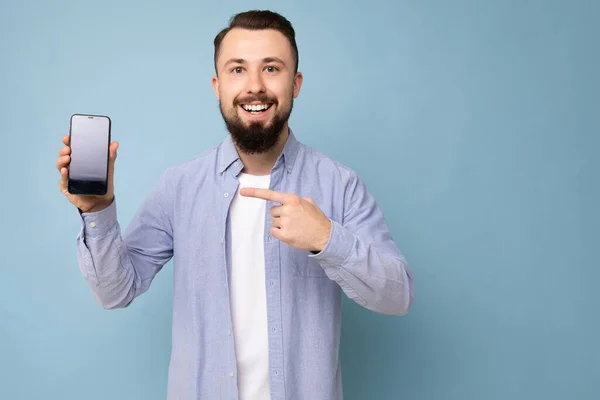 Foto de hombre adulto guapo sonriente guapo usando atuendo casual de pie aislado en el fondo con espacio de copia que sostiene el teléfono inteligente que muestra el teléfono en la mano con pantalla vacía para —  Fotos de Stock