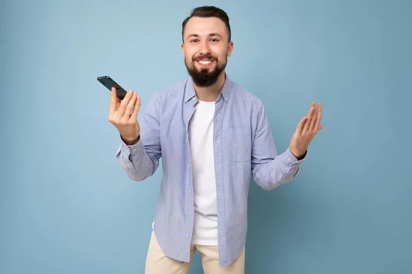 Delightful handsome young brunette unshaven man with beard wearing stylish white t-shirt and blue shirt isolated over blue background with empty space holding in hand and using phone messaging sms — Stock Photo, Image