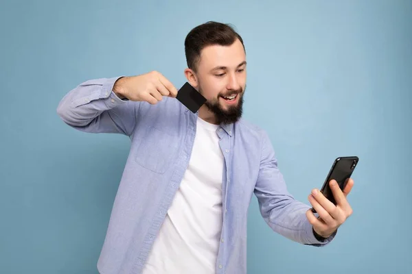 Hombre guapo que usa ropa cotidiana aislada en la pared de fondo sosteniendo y usando el teléfono y la tarjeta de crédito haciendo el pago mirando la pantalla del teléfono inteligente —  Fotos de Stock