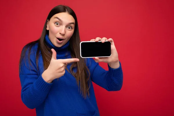 Foto van geschokt vrij gelukkig jonge brunette vrouw dragen casual blauwe trui geïsoleerd over rode achtergrond met lege ruimte in de hand mobiele telefoon en toont smartphone met leeg scherm voor — Stockfoto