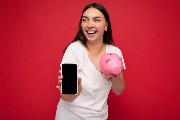 Krásná šťastná mladá brunetka žena na sobě ležérní bílé tričko izolované přes červené pozadí s prázdným prostorem drží v ruce mobilní telefon a ukazuje smartphone s prázdnou obrazovkou pro mockup a — Stock fotografie