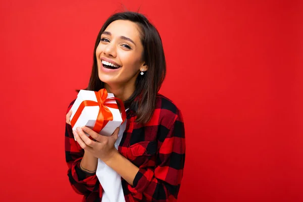Shot of attractive positive smiling young brunette woman isolated over colourful background wall wearing everyday trendy outfit holding gift box and looking at camera — Stock Photo, Image