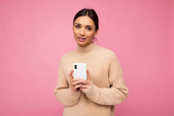 Vonzó aranyos fiatal barna nő visel bézs meleg pulóver áll elszigetelt felett rózsaszín háttér szörfözés az interneten telefonon keresztül nézi a kamerát — Stock Fotó
