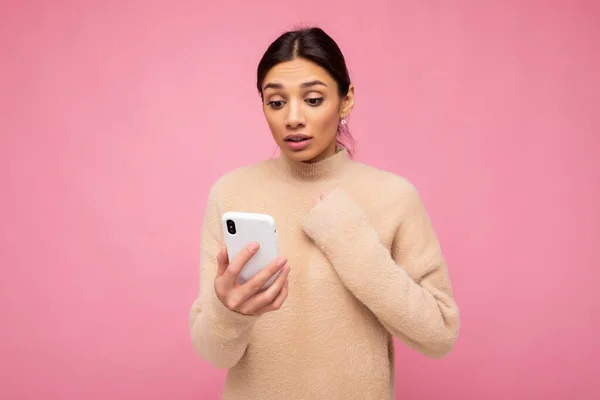 Vonzó aranyos fiatal megdöbbent barna nő visel bézs meleg pulóver elszigetelt felett rózsaszín háttér szörfözés az interneten telefonon keresztül nézi a mobil képernyőn, és tartsa a kezét a szív — Stock Fotó