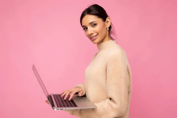 Profil lateral fotografie de frumos zâmbitor tânăr păr întunecat femeie care deține laptop izolat peste fundal de perete uita-te la camera purtând pulover bej — Fotografie, imagine de stoc