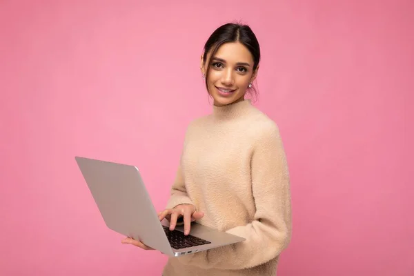 Fotografie de frumoasă tânără brunetă femeie purtând pulover bej care deține netbook calculator tastarea text pe tastatură uitându-se la camera izolată peste fundal roz — Fotografie, imagine de stoc