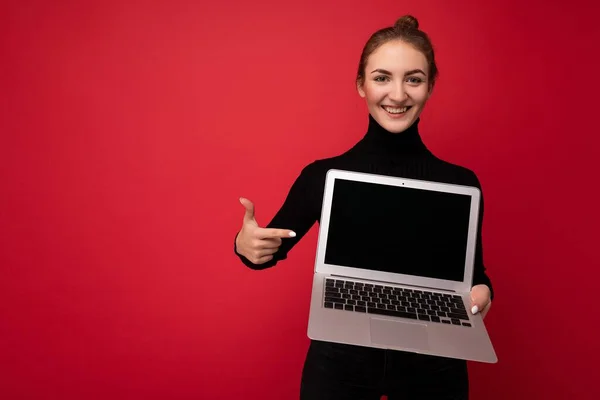 Foto de Hermosa sonriente joven morena feliz sosteniendo computadora portátil con pantalla de monitor vacía con manga larga negro mirando a la cámara pintando en netbook aislado sobre fondo de pared roja —  Fotos de Stock