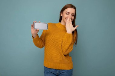 Gülen, turuncu süveter giyen, mavi arka planda yalnız bırakılmış, elinde boş bir alan olan ve cep telefonuna boş bir maket alanı gösteren çekici, pozitif bir genç kadın.