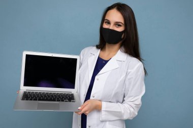 Siyah maskeli, beyaz önlüklü genç bir kadın dizüstü bilgisayar tutuyor ve mavi arka planda izole edilmiş kameraya bakıyor.
