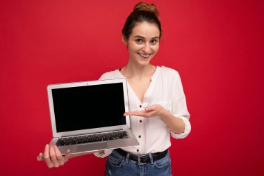 Güzel gülümseyen mutlu genç bir kadının yakın plan portresi. Bilgisayara bakıp kameraya bakıyor. Kırmızı arka planda izole edilmiş şık kıyafetler giyip eğleniyor.