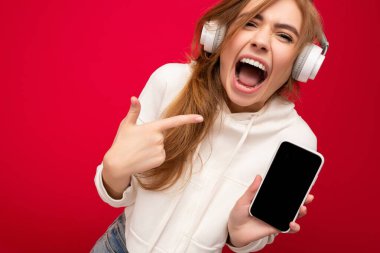 Güzel, şoke olmuş, şık kıyafetler giyen, arka plandaki duvarda izole edilmiş, cep telefonlu, beyaz bluetooth kulaklık takan genç bir kadın.