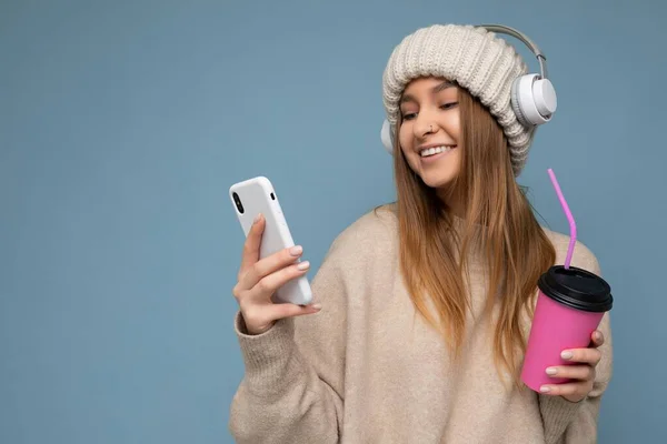 베지 스웨터를 입고 하얀 헤드폰을 낀 매력적으로 웃고 있는 젊은 금발 여성은 손에 파란 배경을 드러내고 휴대 전화를 사용하여 뉴스 음료와 — 스톡 사진