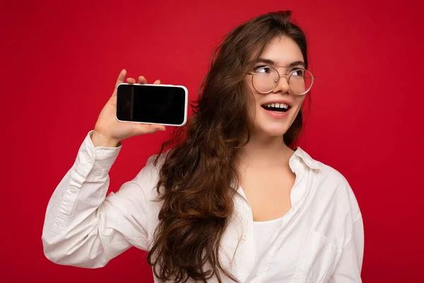 Positivo atraente jovem morena vestindo camisa branca e óculos ópticos isolados sobre fundo vermelho segurando na mão e mostrando telefone celular com tela vazia para mockup olhando para o lado — Fotografia de Stock