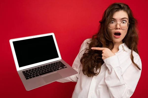 Shocked amazed beautiful brunet curly young woman saying wow holding computer laptop wearing glasses white shirt looking at camera isolated over red wall background