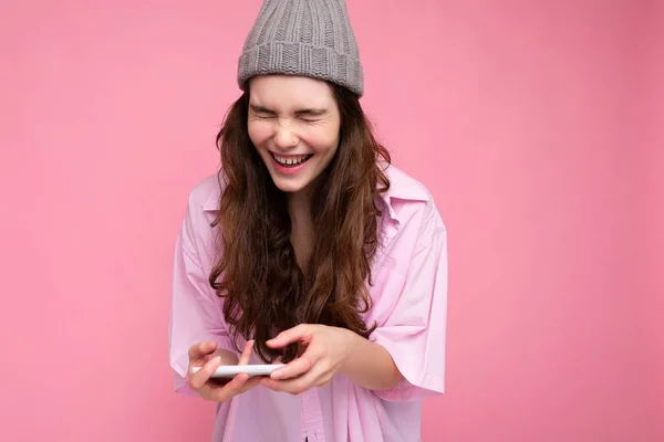 Lachend attraktive positive junge brünette Frau trägt stilvolles rosa Hemd und grauen Hut isoliert über rosa Hintergrund hält in der Hand und mit dem Handy Online-Spiele spielen — Stockfoto