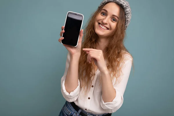 단정 치 못한 흰색 셔츠와 회색 모자를 착용하고 전화기를 들고 있는 푸른 배경 벽에 고립 된 아름다운 금발의 젊은 여성의 사진을 클로즈업 — 스톡 사진