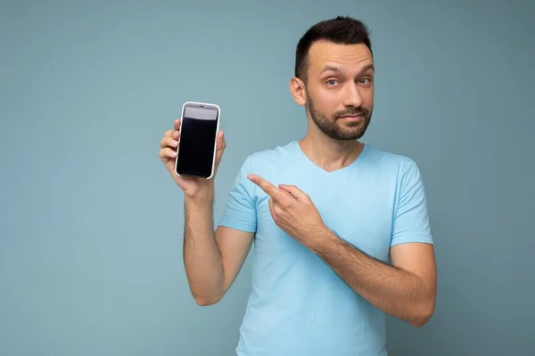 잘생긴 젊고 면도를 하지 않은 브라 운트 셔츠를 입고 파란 배경 위에 고립 된 채 카메라와 손가락을 가리키며 빈 디스플레이가 달린 휴대 전화를 보여 주고 있다. — 스톡 사진