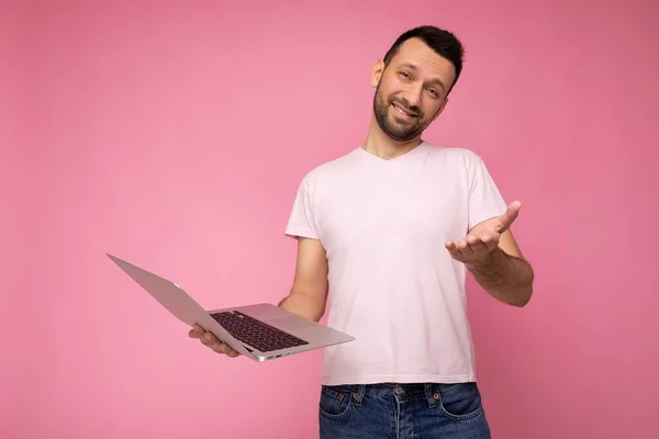Guapo divertido y sonriente morena hombre sosteniendo ordenador portátil y mostrando la mano mirando a la cámara en camiseta sobre fondo rosa aislado — Foto de Stock