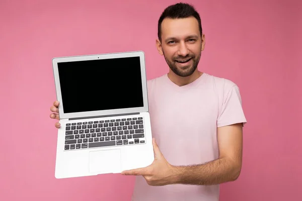 Hombre guapo sosteniendo ordenador portátil mirando a la cámara en camiseta sobre fondo rosa aislado —  Fotos de Stock