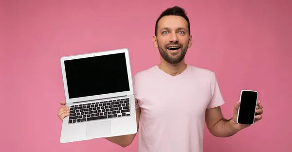 英俊而惊奇的黑发男人拿着手提电脑和手机，穿着孤立的粉色背景T恤看着相机 — 图库照片