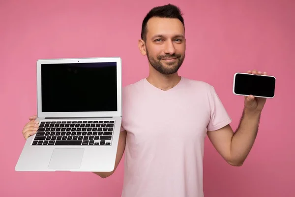 Hombre guapo morena sosteniendo ordenador portátil y teléfono móvil mirando a la cámara en camiseta sobre fondo rosa aislado —  Fotos de Stock